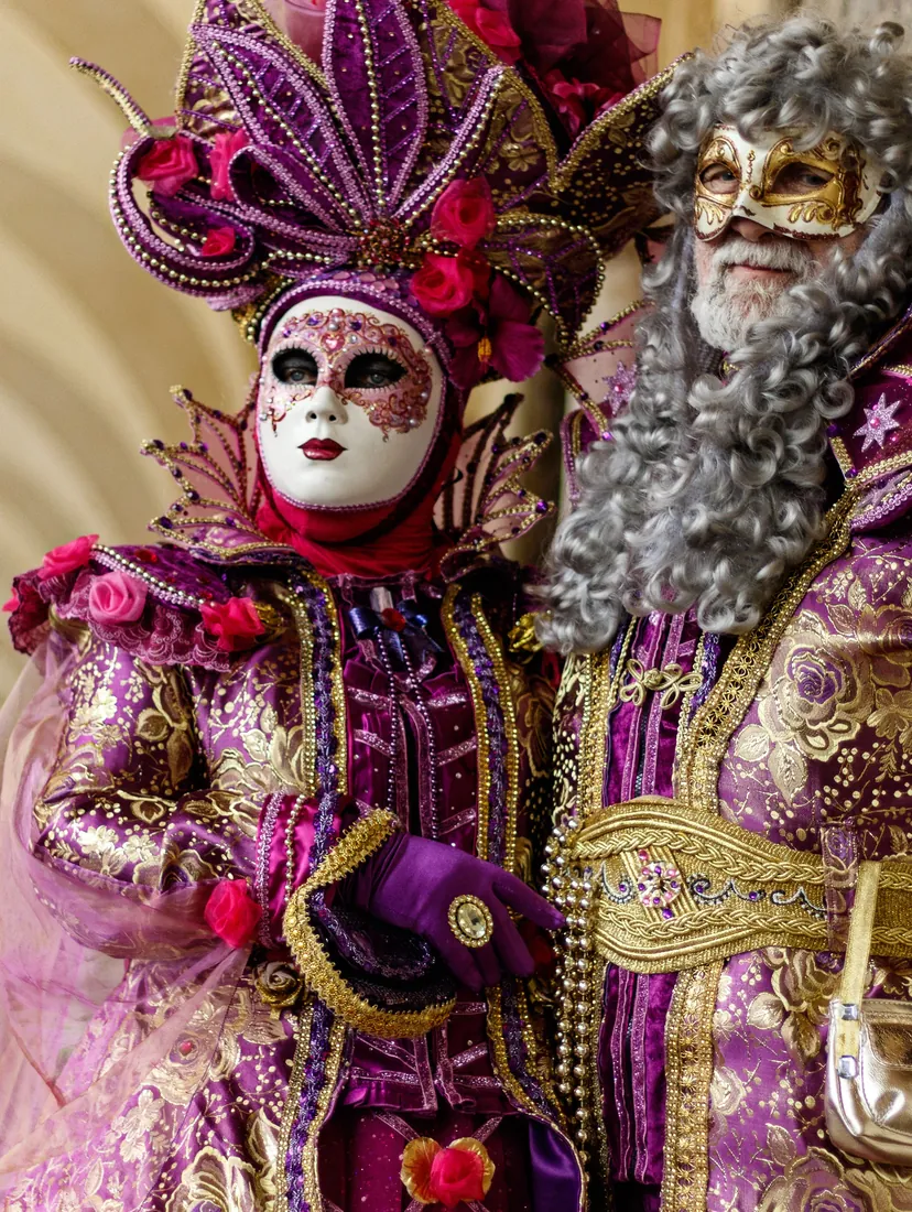 A woman in a purple and gold costume with a rose-trimmed headdress and a white mask beside a man in a grey wig and a gold half-mask, under a stone arcade
