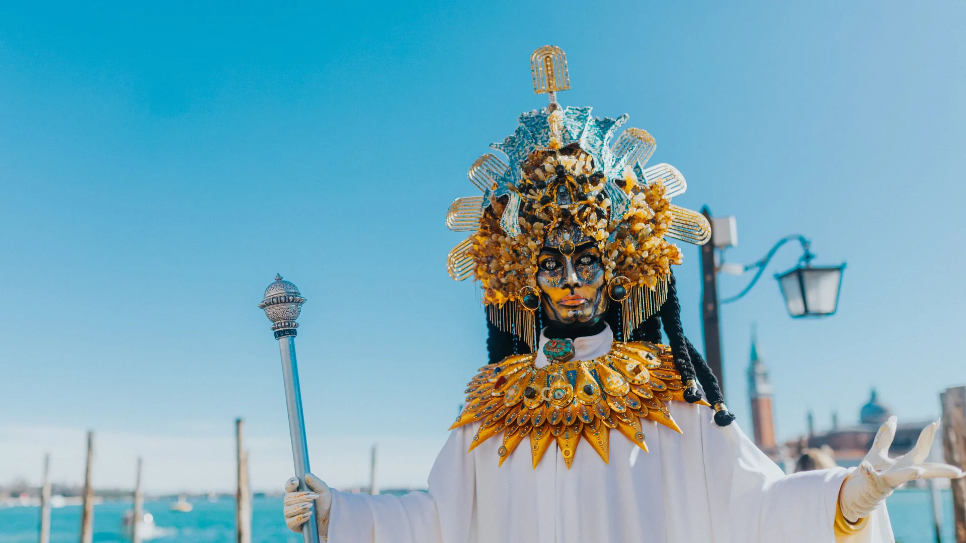 A masked figure in a gold headdress, gold collar and white robes with a silver sceptre, on the Venice waterfront with mooring poles and the lagoon behind