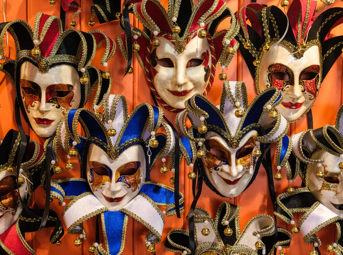 A wall of jester masks in black, gold, red and blue with bells, hung against an orange background